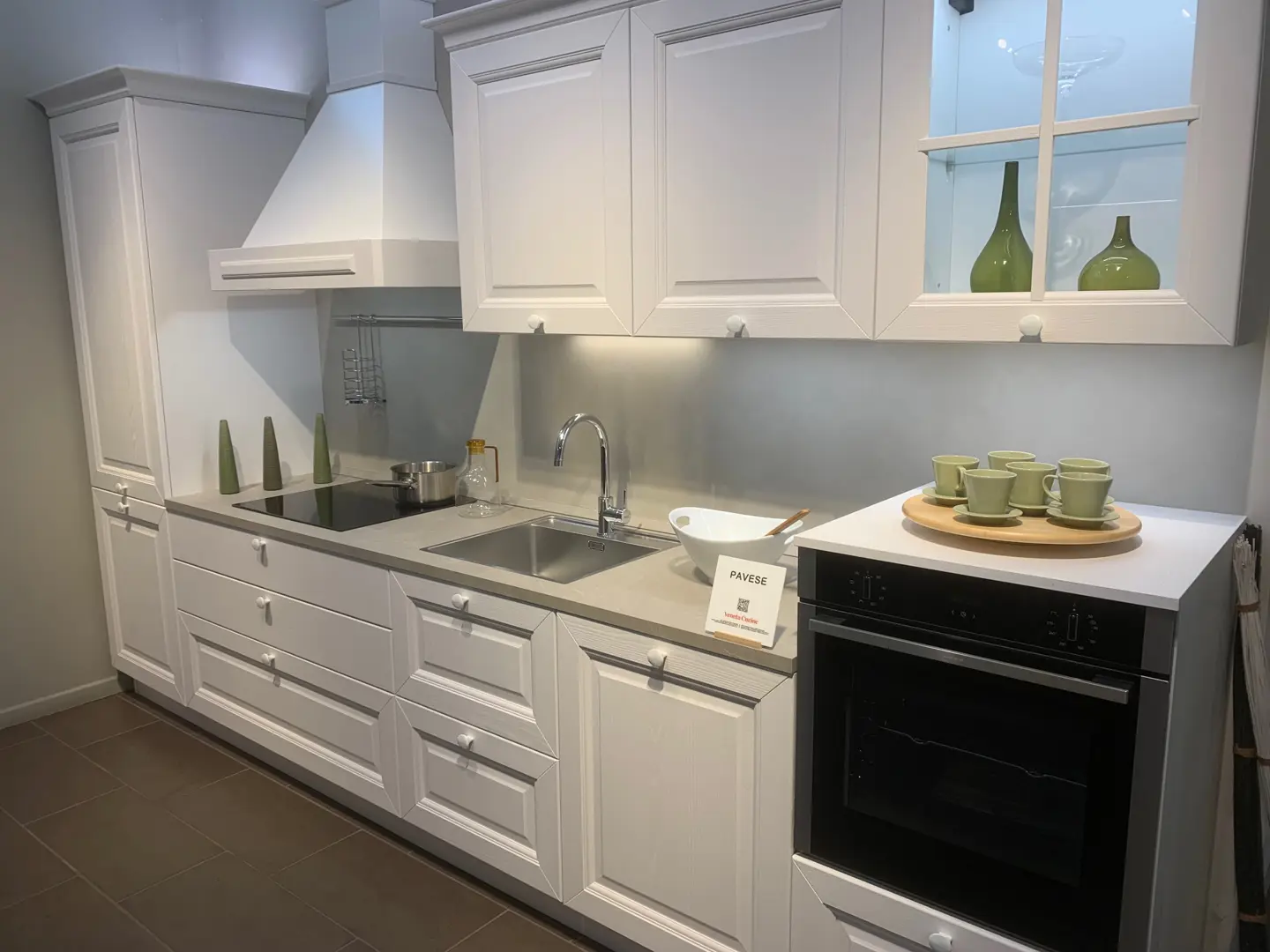Cucina Pavese di Veneta Cucine in frassino laccato bianco con ante a telaio, cappa a camino, piano cottura a induzione e forno a colonna, esposta nello showroom Arredamenti Soldi a Follonica